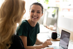 Zwei junge Frauen im Gespräch an einem Schreibtisch mit Laptop, eine lächelt freundlich