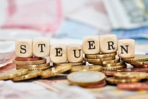 Holzwürfel mit der Aufschrift „STEUERN" auf Münzstapeln und Euroscheinen